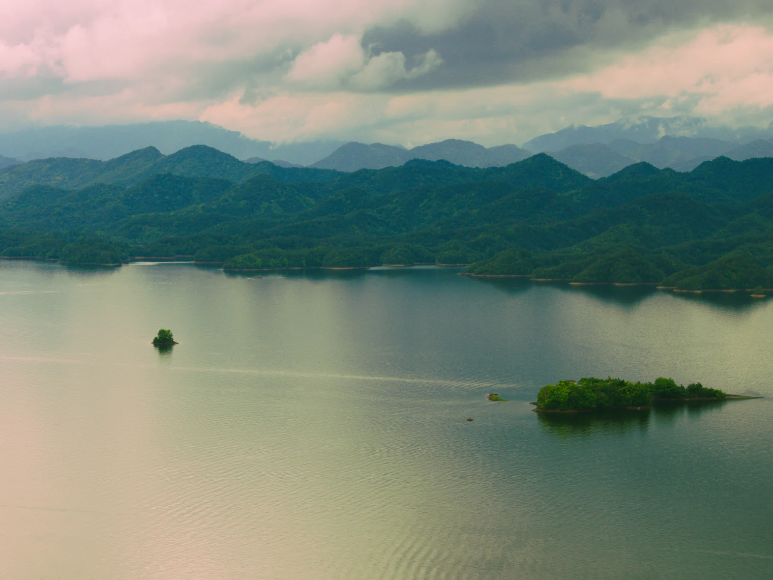 云层下的千岛湖全景，远山层叠，两座小岛浮在平静的湖面上。